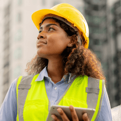 Woman wearing safety vest and hardhat holding a computer tablet looking up at a building.